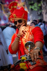 A man dressed in traditional attire, featuring a vibrant red turban and sherwani with gold accents, is posing with a thoughtful expression while sitting on a motorbike. He is wearing sunglasses and has a beard, surrounded by a lively background with people and trees.
