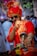A man dressed in traditional attire, featuring a vibrant red turban and sherwani with gold accents, is posing with a thoughtful expression while sitting on a motorbike. He is wearing sunglasses and has a beard, surrounded by a lively background with people and trees.