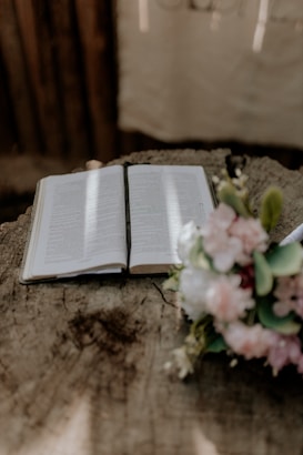 An open book lies on a textured wooden surface, with soft light streaming across its pages. To the right, a bouquet of pink and white flowers is visible, adding a touch of color and natural elegance. The setting suggests a peaceful, contemplative atmosphere.