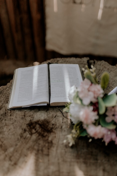 An open book lies on a textured wooden surface, with soft light streaming across its pages. To the right, a bouquet of pink and white flowers is visible, adding a touch of color and natural elegance. The setting suggests a peaceful, contemplative atmosphere.
