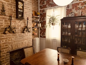 A cozy dining room with a rustic brick wall and vintage decor. The wall features a classic clock and ornate candle holders, flanked by a wooden shelf with culinary items like pots and baskets. A plant hangs from the ceiling in front of a window with sheer white curtains. To the right, a large wooden cabinet displays dinnerware. The table in the foreground has two chairs and is adorned with small bottles and a dish.