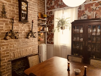 A cozy dining room with a rustic brick wall and vintage decor. The wall features a classic clock and ornate candle holders, flanked by a wooden shelf with culinary items like pots and baskets. A plant hangs from the ceiling in front of a window with sheer white curtains. To the right, a large wooden cabinet displays dinnerware. The table in the foreground has two chairs and is adorned with small bottles and a dish.