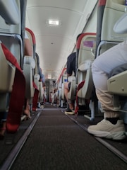 A view from the aisle of an airplane cabin showing rows of seats with passengers sitting. The seats have red accents and visible seatbelts. Overhead compartments are present, and there is an exit sign at the top.