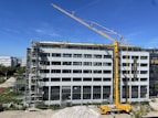 A large construction site with a modern multi-story building under renovation. A yellow crane is positioned near the building, and several workers in safety gear are visible on the rooftop, indicating ongoing construction work. Scaffolding is set up along the side of the building. The scene is sunlit under a clear blue sky.