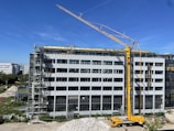 A large construction site with a modern multi-story building under renovation. A yellow crane is positioned near the building, and several workers in safety gear are visible on the rooftop, indicating ongoing construction work. Scaffolding is set up along the side of the building. The scene is sunlit under a clear blue sky.