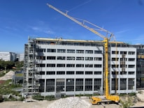 A large construction site with a modern multi-story building under renovation. A yellow crane is positioned near the building, and several workers in safety gear are visible on the rooftop, indicating ongoing construction work. Scaffolding is set up along the side of the building. The scene is sunlit under a clear blue sky.