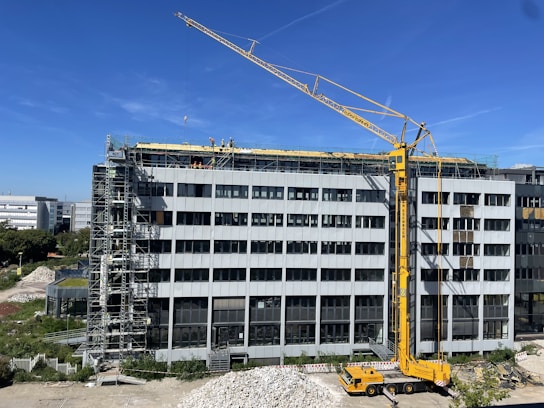 A large construction site with a modern multi-story building under renovation. A yellow crane is positioned near the building, and several workers in safety gear are visible on the rooftop, indicating ongoing construction work. Scaffolding is set up along the side of the building. The scene is sunlit under a clear blue sky.