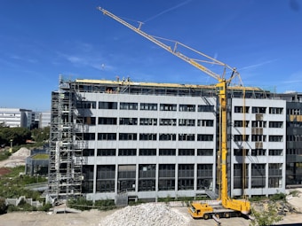 A large construction site with a modern multi-story building under renovation. A yellow crane is positioned near the building, and several workers in safety gear are visible on the rooftop, indicating ongoing construction work. Scaffolding is set up along the side of the building. The scene is sunlit under a clear blue sky.