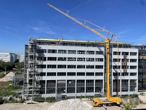 A large construction site with a modern multi-story building under renovation. A yellow crane is positioned near the building, and several workers in safety gear are visible on the rooftop, indicating ongoing construction work. Scaffolding is set up along the side of the building. The scene is sunlit under a clear blue sky.