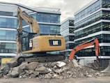 A construction site featuring two excavators, one yellow and one orange, positioned on a pile of broken concrete and rocks. Modern office buildings with glass facades form the backdrop. The scene indicates active demolition or construction activity.