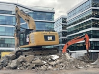 A construction site featuring two excavators, one yellow and one orange, positioned on a pile of broken concrete and rocks. Modern office buildings with glass facades form the backdrop. The scene indicates active demolition or construction activity.