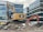 A construction site featuring two excavators, one yellow and one orange, positioned on a pile of broken concrete and rocks. Modern office buildings with glass facades form the backdrop. The scene indicates active demolition or construction activity.