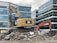 A construction site featuring two excavators, one yellow and one orange, positioned on a pile of broken concrete and rocks. Modern office buildings with glass facades form the backdrop. The scene indicates active demolition or construction activity.