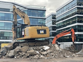 A construction site featuring two excavators, one yellow and one orange, positioned on a pile of broken concrete and rocks. Modern office buildings with glass facades form the backdrop. The scene indicates active demolition or construction activity.