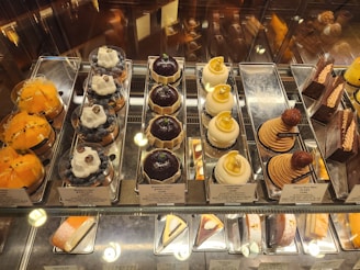 An assortment of gourmet pastries displayed in a glass case. There are several types including yellow fruit tarts, blueberry tarts topped with whipped cream and a blueberry, domed chocolate pastries, domed coconut pastries with gold leaf pieces, stack-shaped pastries with a chestnut on top, and various chocolate cakes. Each item is neatly arranged on silver trays with small signage indicating the name and price.