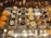 An assortment of gourmet pastries displayed in a glass case. There are several types including yellow fruit tarts, blueberry tarts topped with whipped cream and a blueberry, domed chocolate pastries, domed coconut pastries with gold leaf pieces, stack-shaped pastries with a chestnut on top, and various chocolate cakes. Each item is neatly arranged on silver trays with small signage indicating the name and price.