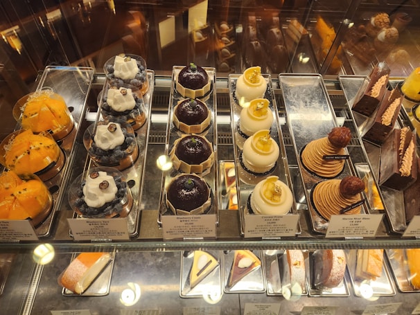 An assortment of gourmet pastries displayed in a glass case. There are several types including yellow fruit tarts, blueberry tarts topped with whipped cream and a blueberry, domed chocolate pastries, domed coconut pastries with gold leaf pieces, stack-shaped pastries with a chestnut on top, and various chocolate cakes. Each item is neatly arranged on silver trays with small signage indicating the name and price.