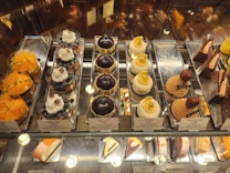 An assortment of gourmet pastries displayed in a glass case. There are several types including yellow fruit tarts, blueberry tarts topped with whipped cream and a blueberry, domed chocolate pastries, domed coconut pastries with gold leaf pieces, stack-shaped pastries with a chestnut on top, and various chocolate cakes. Each item is neatly arranged on silver trays with small signage indicating the name and price.