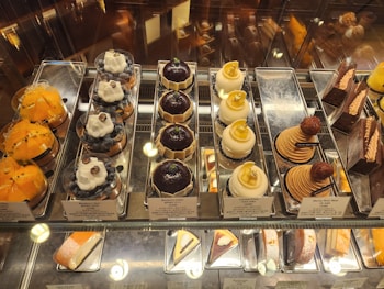 An assortment of gourmet pastries displayed in a glass case. There are several types including yellow fruit tarts, blueberry tarts topped with whipped cream and a blueberry, domed chocolate pastries, domed coconut pastries with gold leaf pieces, stack-shaped pastries with a chestnut on top, and various chocolate cakes. Each item is neatly arranged on silver trays with small signage indicating the name and price.