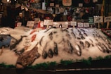 A bustling fish market stall displays a variety of seafood, including several whole fish on a bed of crushed ice. Handwritten signs indicate the types and prices of the fish, such as monkfish and halibut. The market ambiance is lively, with customers and vendors in the background. The environment is dimly lit with a warm, inviting atmosphere.