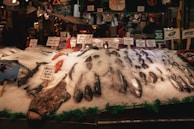 A bustling fish market stall displays a variety of seafood, including several whole fish on a bed of crushed ice. Handwritten signs indicate the types and prices of the fish, such as monkfish and halibut. The market ambiance is lively, with customers and vendors in the background. The environment is dimly lit with a warm, inviting atmosphere.