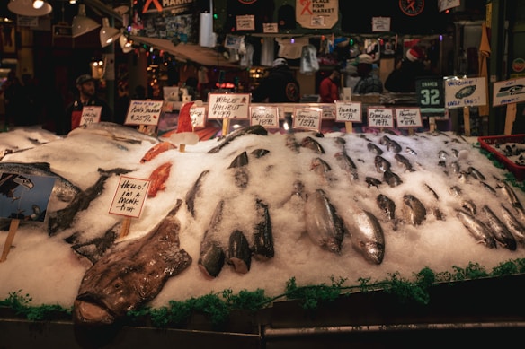 A bustling fish market stall displays a variety of seafood, including several whole fish on a bed of crushed ice. Handwritten signs indicate the types and prices of the fish, such as monkfish and halibut. The market ambiance is lively, with customers and vendors in the background. The environment is dimly lit with a warm, inviting atmosphere.