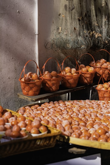 Numerous brown eggs are displayed in orange baskets and large woven trays, with the eggs arranged in a market setting. The background includes a textured wall and fishing nets draped above the baskets, suggesting a rustic or artisanal environment.
