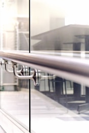 A close-up view of a modern railing in a building, with a sleek stainless steel design and glass panels. The background appears to be an office or a public space with tables and chairs.
