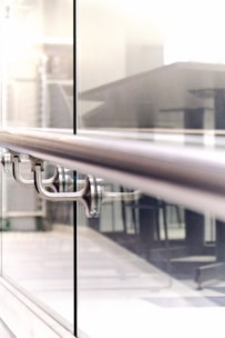 A close-up view of a modern railing in a building, with a sleek stainless steel design and glass panels. The background appears to be an office or a public space with tables and chairs.