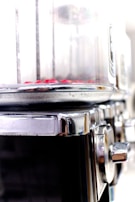 A close-up view of a metallic cylindrical appliance with a transparent section at the top containing small red objects. The appliance has a shiny, reflective metal surface with a knob on the side, suggesting it might be used for cooking or processing food.