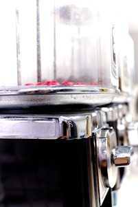 A close-up view of a metallic cylindrical appliance with a transparent section at the top containing small red objects. The appliance has a shiny, reflective metal surface with a knob on the side, suggesting it might be used for cooking or processing food.