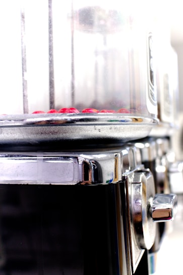 A close-up view of a metallic cylindrical appliance with a transparent section at the top containing small red objects. The appliance has a shiny, reflective metal surface with a knob on the side, suggesting it might be used for cooking or processing food.