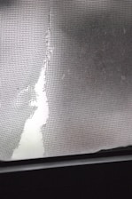 A torn and damaged wire mesh screen is lit by soft light, revealing a large tear. The jagged edges of the screen contrast with a smooth, blurred background.