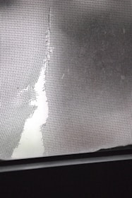 A torn and damaged wire mesh screen is lit by soft light, revealing a large tear. The jagged edges of the screen contrast with a smooth, blurred background.