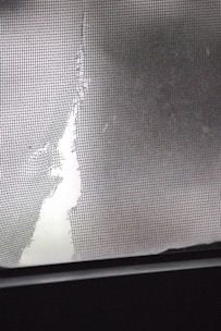 A torn and damaged wire mesh screen is lit by soft light, revealing a large tear. The jagged edges of the screen contrast with a smooth, blurred background.