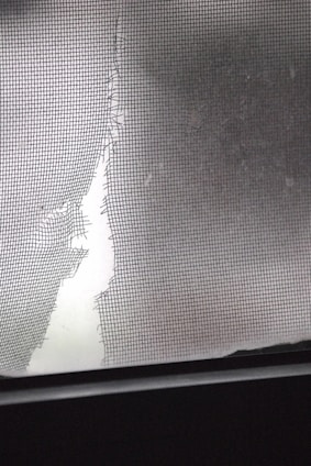 A torn and damaged wire mesh screen is lit by soft light, revealing a large tear. The jagged edges of the screen contrast with a smooth, blurred background.