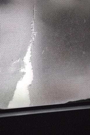 A torn and damaged wire mesh screen is lit by soft light, revealing a large tear. The jagged edges of the screen contrast with a smooth, blurred background.