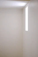 A minimalist empty white room with soft natural light streaming through a large window.