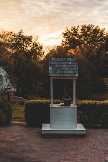 A rustic well with a shingled roof stands on a brick path, surrounded by hedges and trees in warm autumn colors at sunset. A wooden bucket hangs from the well's pulley, creating an old-fashioned and serene scene.