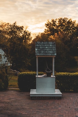 A rustic well with a shingled roof stands on a brick path, surrounded by hedges and trees in warm autumn colors at sunset. A wooden bucket hangs from the well's pulley, creating an old-fashioned and serene scene.