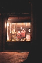 A warmly lit store window displays mannequins dressed in red clothing. Twinkling string lights adorn the lower part of the window, adding a festive touch. The exterior is made of dark brick, and a large tree trunk is visible on the right side. The scene exudes a cozy and inviting ambiance typical of holiday decorations.
