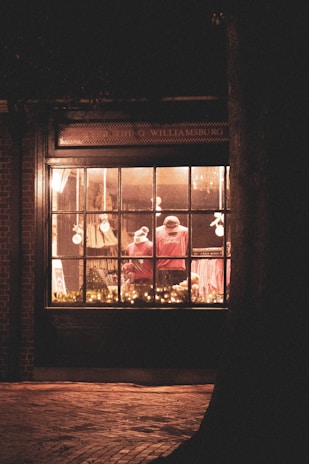 A warmly lit store window displays mannequins dressed in red clothing. Twinkling string lights adorn the lower part of the window, adding a festive touch. The exterior is made of dark brick, and a large tree trunk is visible on the right side. The scene exudes a cozy and inviting ambiance typical of holiday decorations.