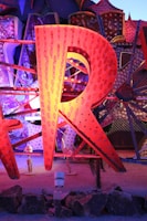 A large, illuminated letter 'R' made from metal, featuring an array of small light bulbs. The letter is part of a larger display with other letters partially visible in the background, all of which are constructed in a similar style. These elements are set against a textured, polka-dotted backdrop with various geometric patterns. The scene is lit with vibrant colors including shades of orange, blue, and purple.
