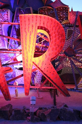 A large, illuminated letter 'R' made from metal, featuring an array of small light bulbs. The letter is part of a larger display with other letters partially visible in the background, all of which are constructed in a similar style. These elements are set against a textured, polka-dotted backdrop with various geometric patterns. The scene is lit with vibrant colors including shades of orange, blue, and purple.