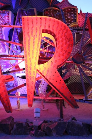 A large, illuminated letter 'R' made from metal, featuring an array of small light bulbs. The letter is part of a larger display with other letters partially visible in the background, all of which are constructed in a similar style. These elements are set against a textured, polka-dotted backdrop with various geometric patterns. The scene is lit with vibrant colors including shades of orange, blue, and purple.