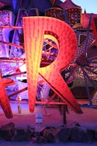 A large, illuminated letter 'R' made from metal, featuring an array of small light bulbs. The letter is part of a larger display with other letters partially visible in the background, all of which are constructed in a similar style. These elements are set against a textured, polka-dotted backdrop with various geometric patterns. The scene is lit with vibrant colors including shades of orange, blue, and purple.