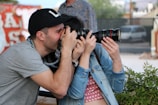 A person wearing a black cap is enthusiastically taking a photograph with a digital camera. Another person is closely engaging with them, holding the lens of the camera. Surroundings include greenery in the foreground and blurred buildings in the background.
