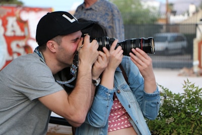 A person wearing a black cap is enthusiastically taking a photograph with a digital camera. Another person is closely engaging with them, holding the lens of the camera. Surroundings include greenery in the foreground and blurred buildings in the background.