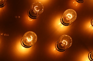 Several light bulbs arranged on a flat surface, each bulb emitting a warm, glowing light. The bulbs' reflections can be seen on the metallic background, enhancing the overall warm and illuminated feel of the scene.