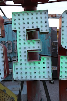 A large illuminated sign featuring a letter 'E', adorned with numerous bulbs, set against a rustic and slightly worn backdrop. The sign exhibits an industrial feel with hints of oxidation and weathering.